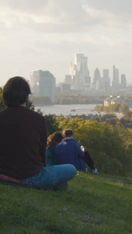 Vertical-Video-Of-People-Sitting-On-Hill-In-Greenwich-Park-Looking-Out-Over-View-Of-City-Skyline-And-River-Thames-In-London-UK